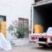 Cardboard boxes and draped chairs sit inside and beside an open truck parked in front of a white house.