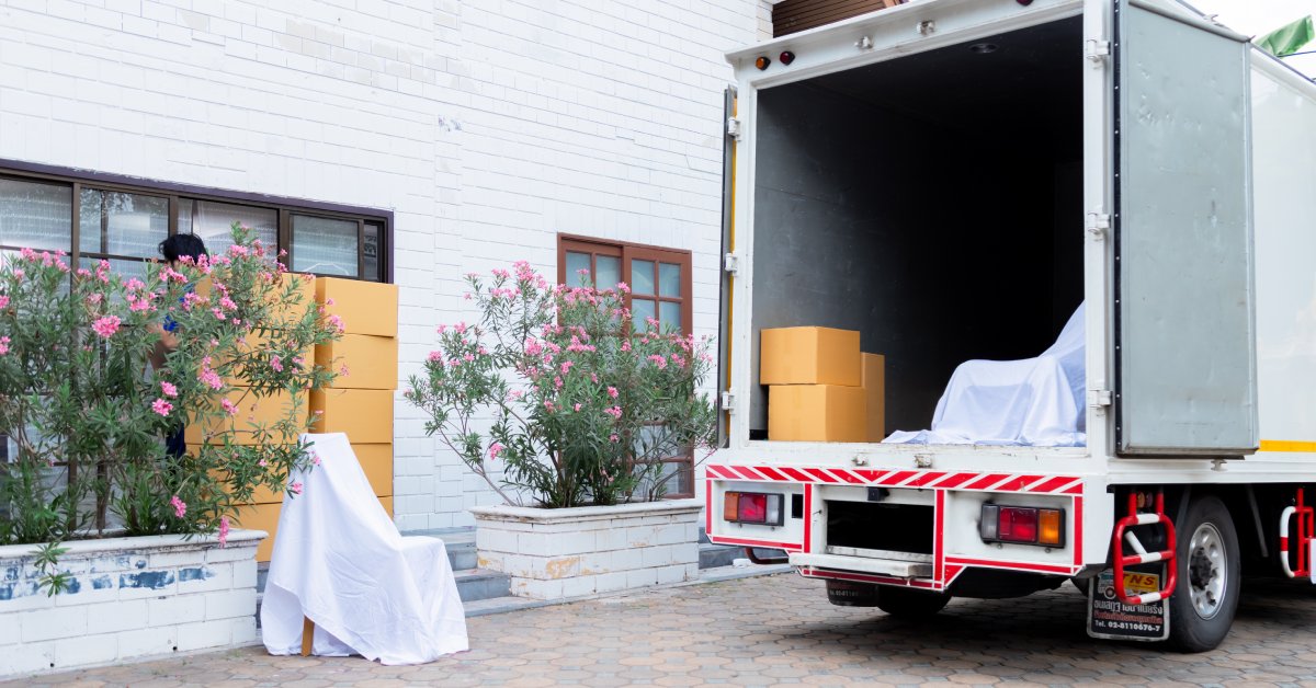 Cardboard boxes and draped chairs sit inside and beside an open truck parked in front of a white house.