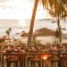 The sun sets on a beach wedding reception. There are tables decorated with flowers, coconuts, and greenery. The ocean's in the background.