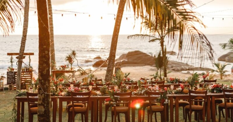 The sun sets on a beach wedding reception. There are tables decorated with flowers, coconuts, and greenery. The ocean's in the background.