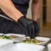 A gloved hand places herbs on food in a Styrofoam box between another box with food and three bowls filled with liquid.