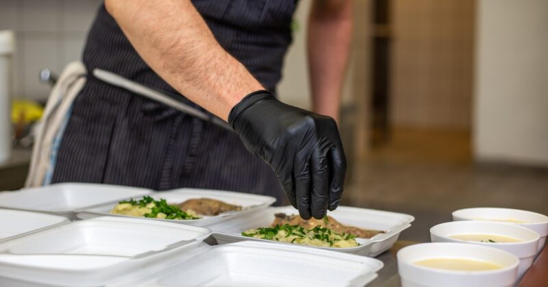 A gloved hand places herbs on food in a Styrofoam box between another box with food and three bowls filled with liquid.