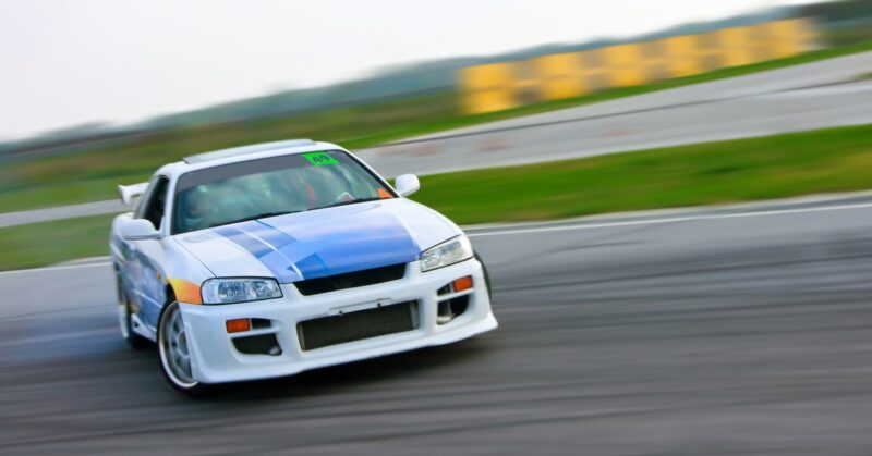 A white race car with a blue stripe and orange details skids along a track, the background blurred from speed.