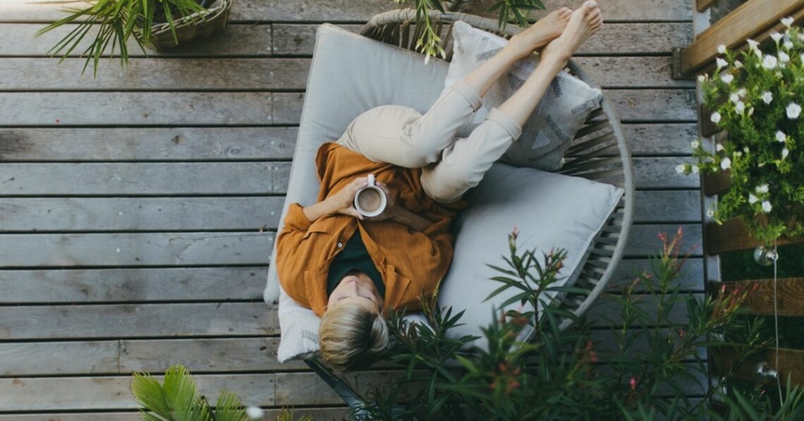A top view of a woman relaxing on her enclosed patio, drinking coffee, and lying on a patio chair, surrounded by plants.