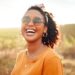 A happy Black woman stands outside in a field in an orange shirt and sunglasses. She's smiling and has a ponytail.