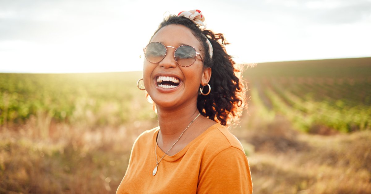 A happy Black woman stands outside in a field in an orange shirt and sunglasses. She's smiling and has a ponytail.