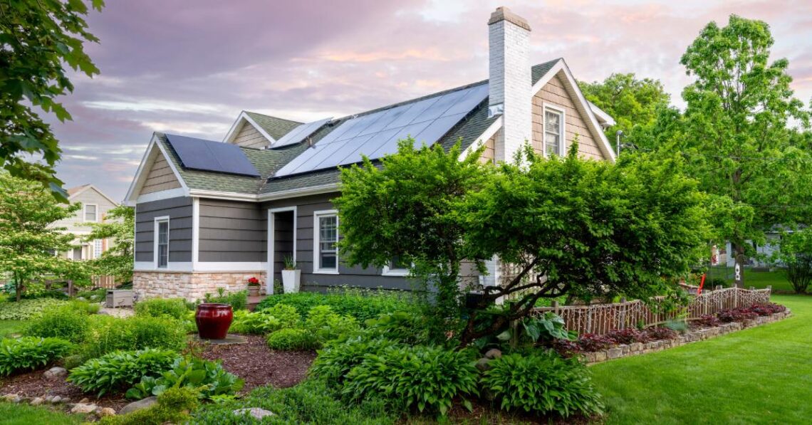 A small house surrounded by luscious trees and green grass. The house's roof is covered with solar panels.