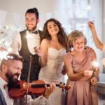 A newlywed couple surrounded by guests. Some hold sparklers, one sings into a microphone, and another plays a violin.Features You Need at Your Wedding Reception
