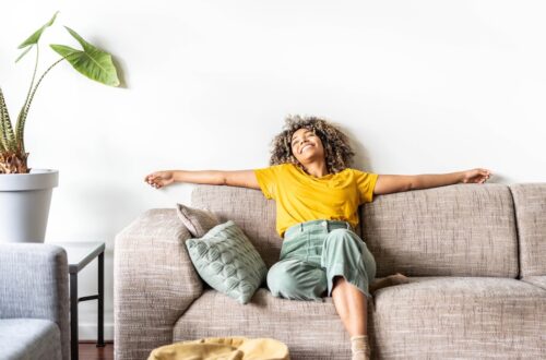 A person wearing a bright yellow shirt is reclining on a couch with their arms spread wide. They are smiling widely.