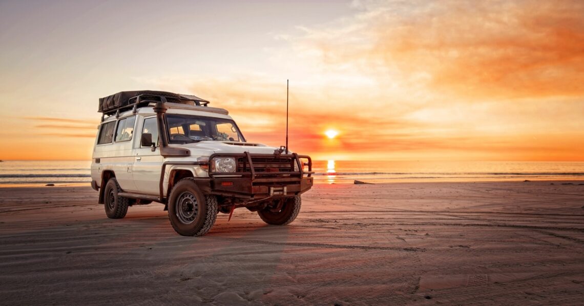A car is parked on the beach with smooth sand and water behind it. The sun is setting in the distance.