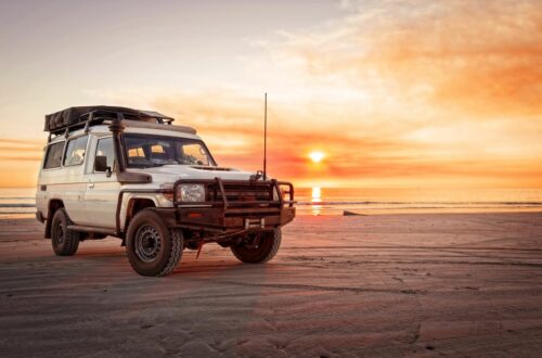 A car is parked on the beach with smooth sand and water behind it. The sun is setting in the distance.