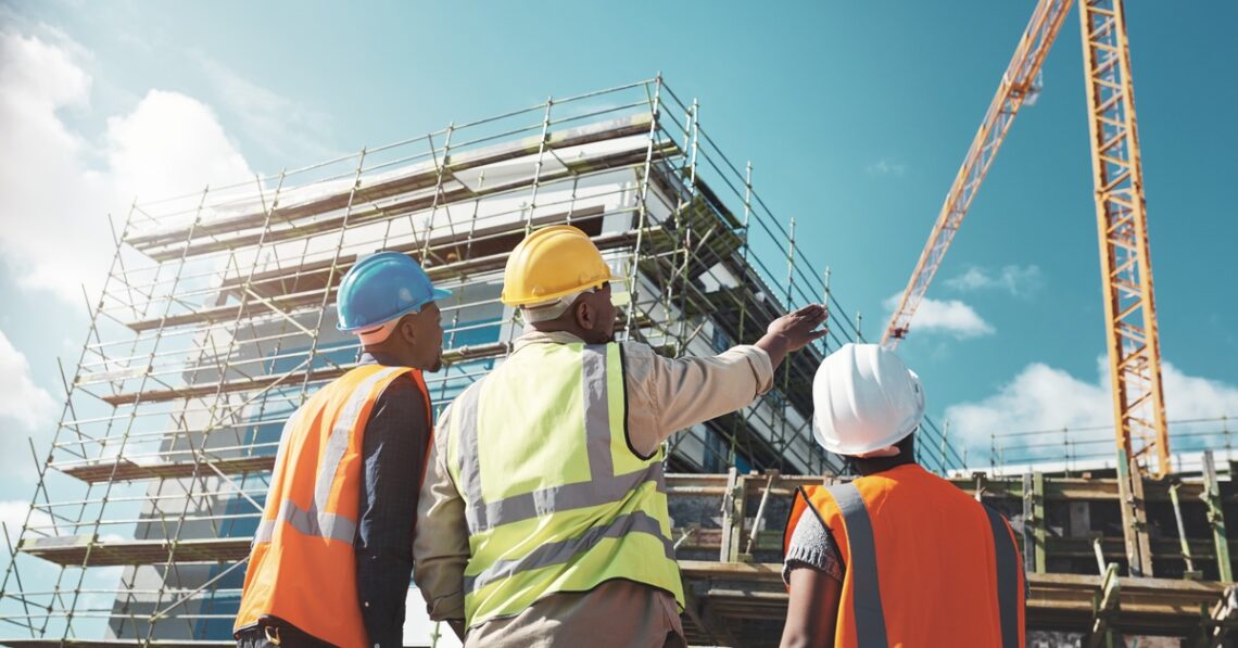 A group of construction workers outside of a building. There is scaffolding and a large crane on the site.