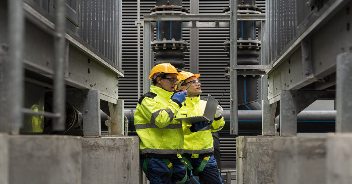 Two industrial engineers examining a piece of equipment. Both of them are wearing high-visibility jackets and helmets.