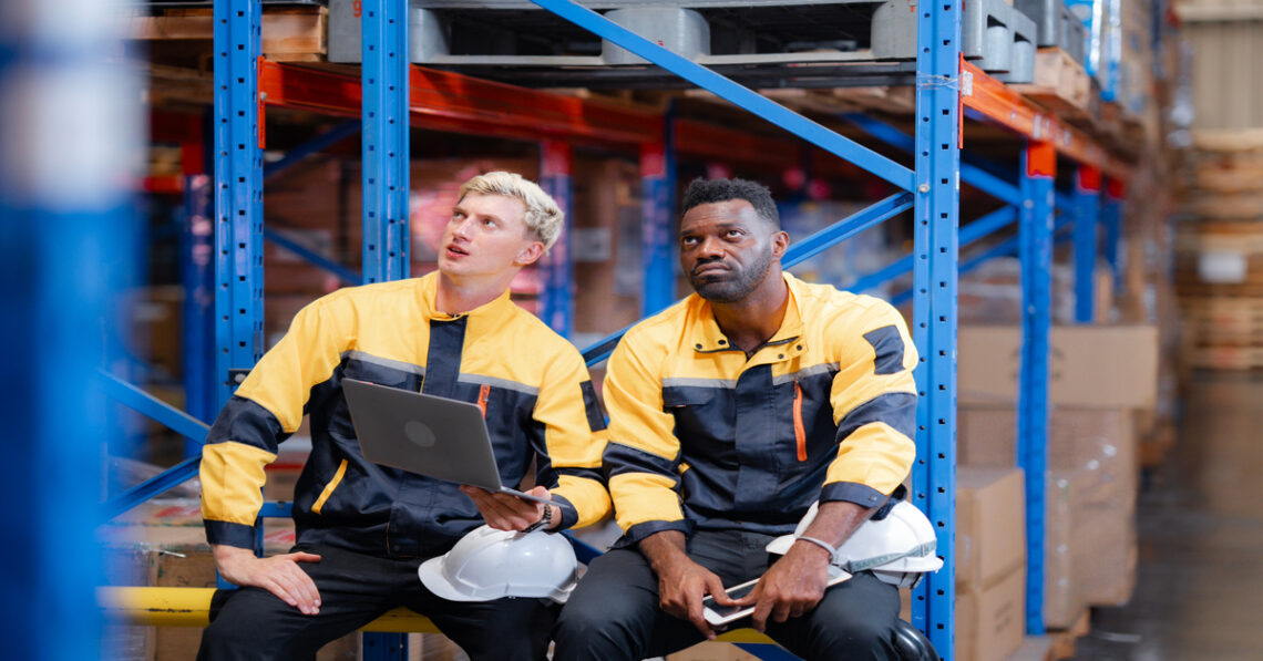 Two coworkers are sitting in a warehouse and looking up at a tall storage shelf. They are both holding safety helmets.