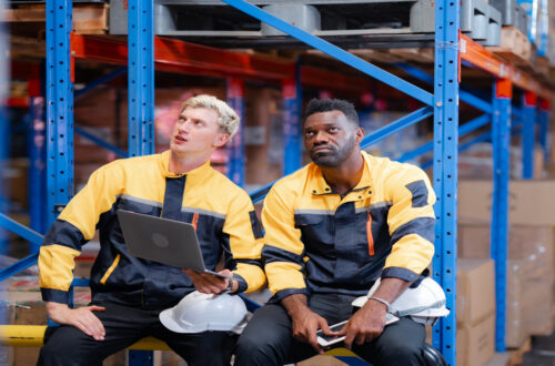 Two coworkers are sitting in a warehouse and looking up at a tall storage shelf. They are both holding safety helmets.