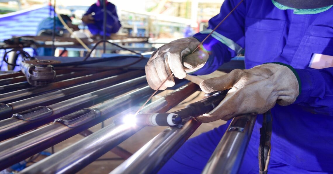 A person in a blue uniform and heat resistant gloves as they work on a piece of metal welding material.