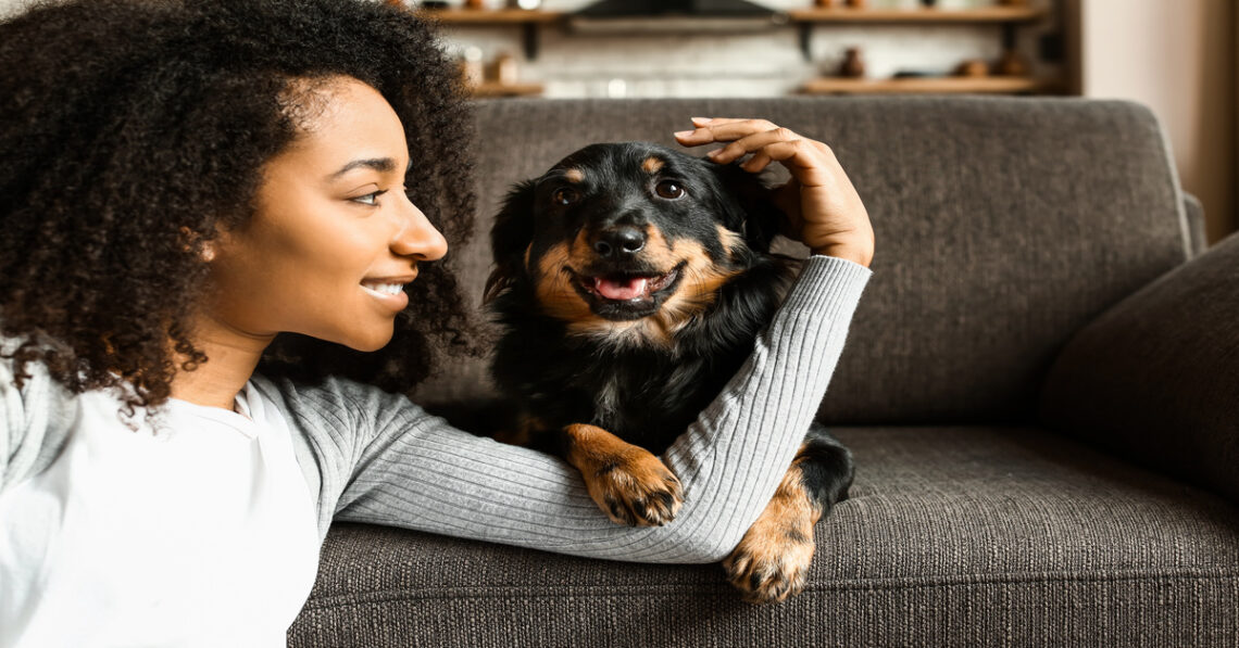 A young Black woman sits on the floor in her home, smiling as she stares at her cute dog on the couch.