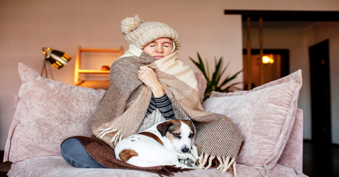 A young woman warms herself up with a cozy hat and a large blanket while sitting on a couch with her dog.