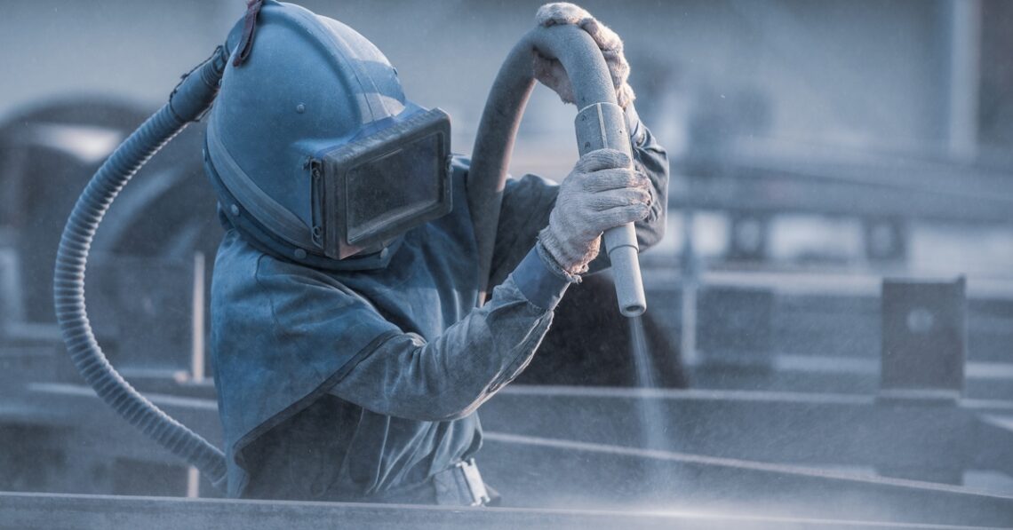 A worker in protective gear operates a sandblasting nozzle, creating a dusty haze in an industrial setting with muted tones.