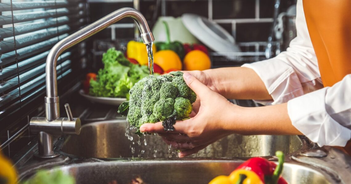 A person is washing some broccoli in the sink. There are other vegetables around the counter.
