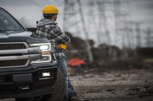 A man in a hard hat is leaning against his car as he’s looking off into the distance at several electrical towers.