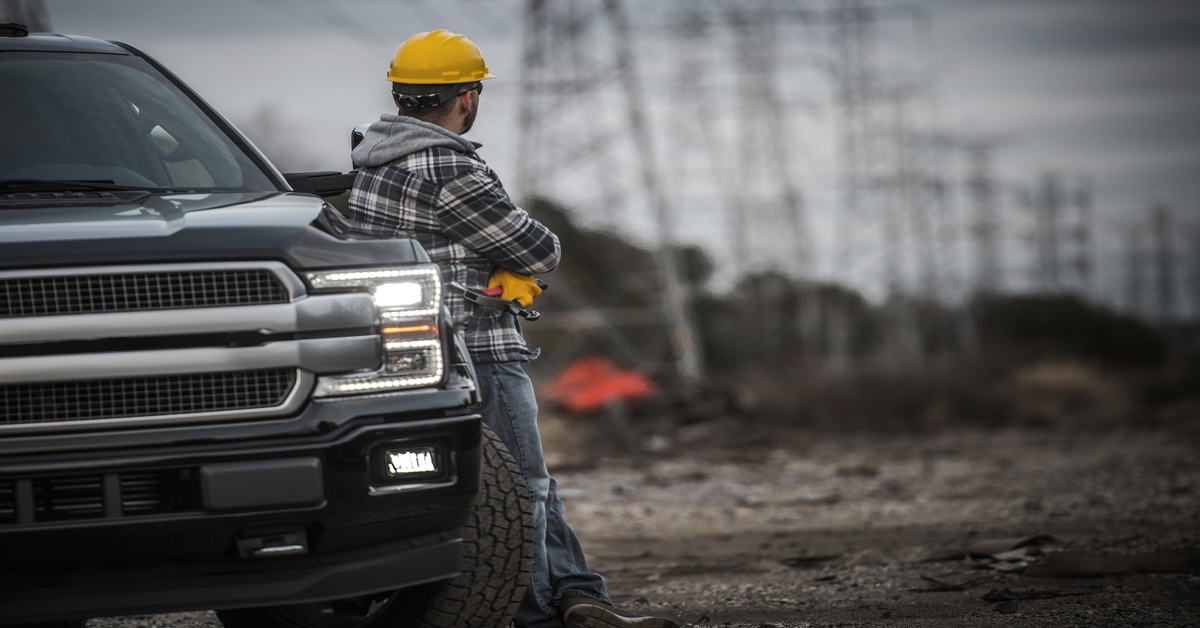 A man in a hard hat is leaning against his car as he’s looking off into the distance at several electrical towers.