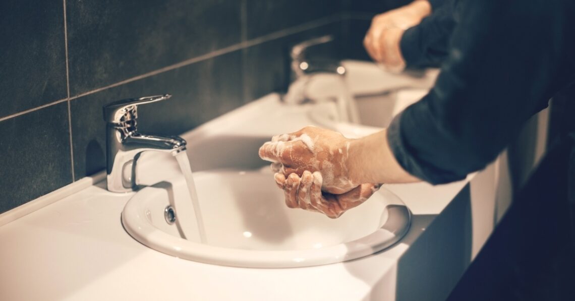 A close-up view shows two people washing their hands in a public restroom. They both have soap on their hands.