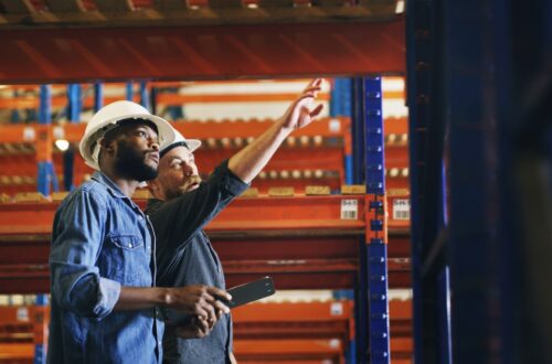 Two construction workers standing in a warehouse and discussing something. One of them is pointing at a pallet rack.
