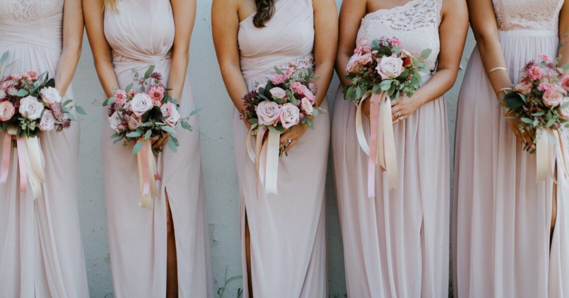 Six bridesmaids in blush pink dresses hold floral bouquets with roses and greenery, standing on grass against a light wall.