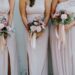 Six bridesmaids in blush pink dresses hold floral bouquets with roses and greenery, standing on grass against a light wall.