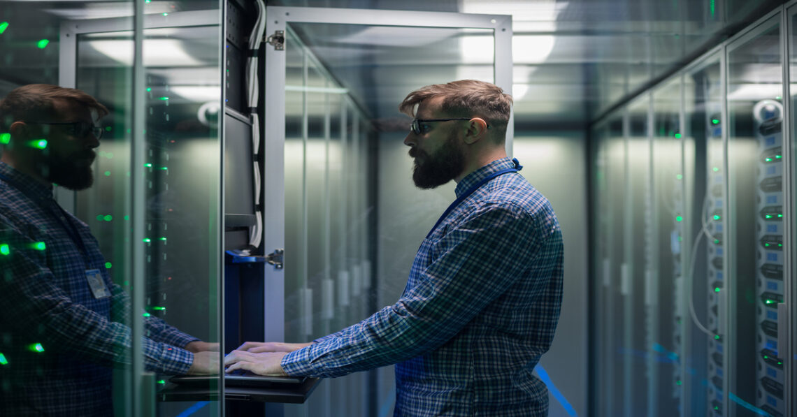 A man with a beard wearing a blue plaid shirt and eyeglasses uses a keyboard on one of the computer server racks.