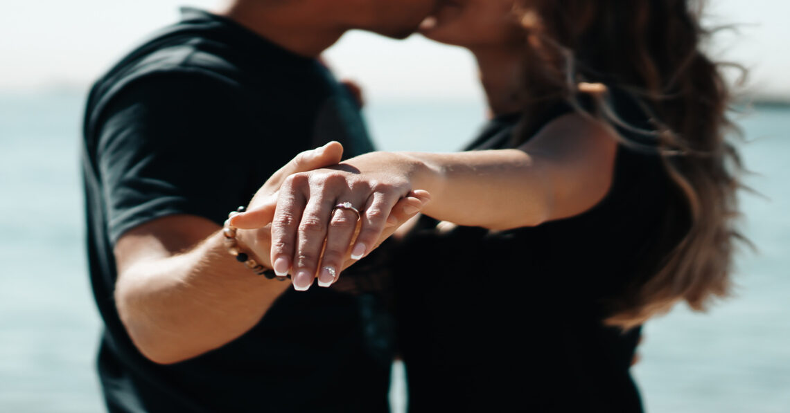 A couple kisses while the man gently holds the woman’s manicured hand toward the viewer, showing her engagement ring.