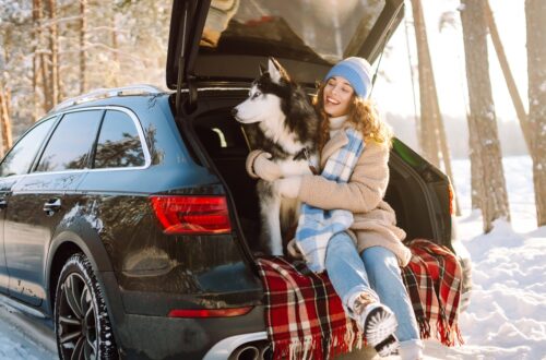 A woman sitting in the trunk of her car with a dog. She is wearing a winter coat and looking at the snow on the ground.