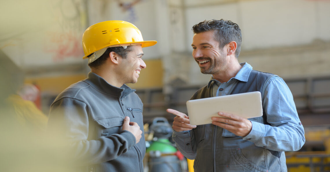 Two men are standing next to each other; one man is wearing a hard hat while the other is not.