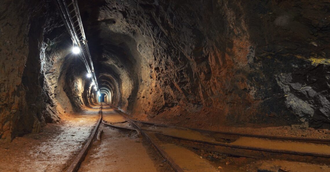 An underground mine features a long passageway with rails for the mine carts. Lights line the top of the passage.