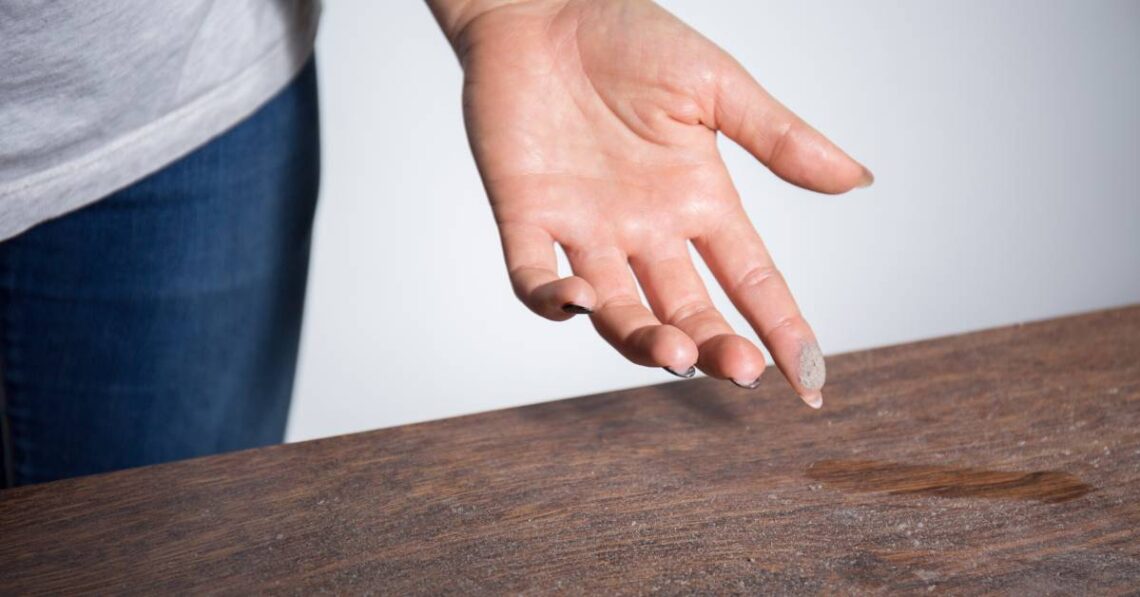 A thin layer of dust is on a wooden table. A person has dust on their fingertip after they swiped the dusty surface.