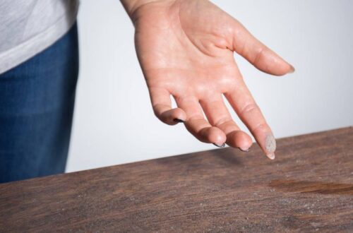 A thin layer of dust is on a wooden table. A person has dust on their fingertip after they swiped the dusty surface.
