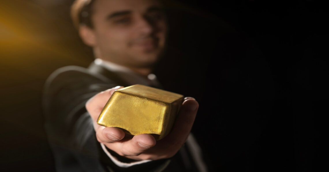 A close-up view shows a person dressed in a suit outstretching their arm to show off a bar of gold.