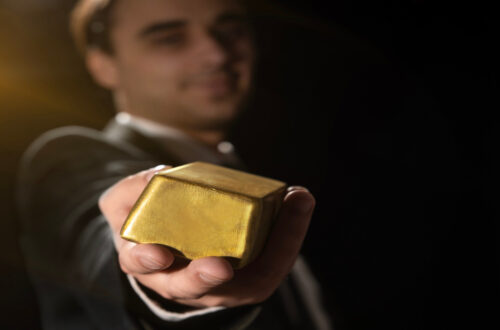 A close-up view shows a person dressed in a suit outstretching their arm to show off a bar of gold.