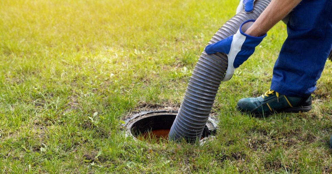 A man in blue gloves feeds a large hose into a septic tank's opening. The opening is in the middle of a grass lawn.