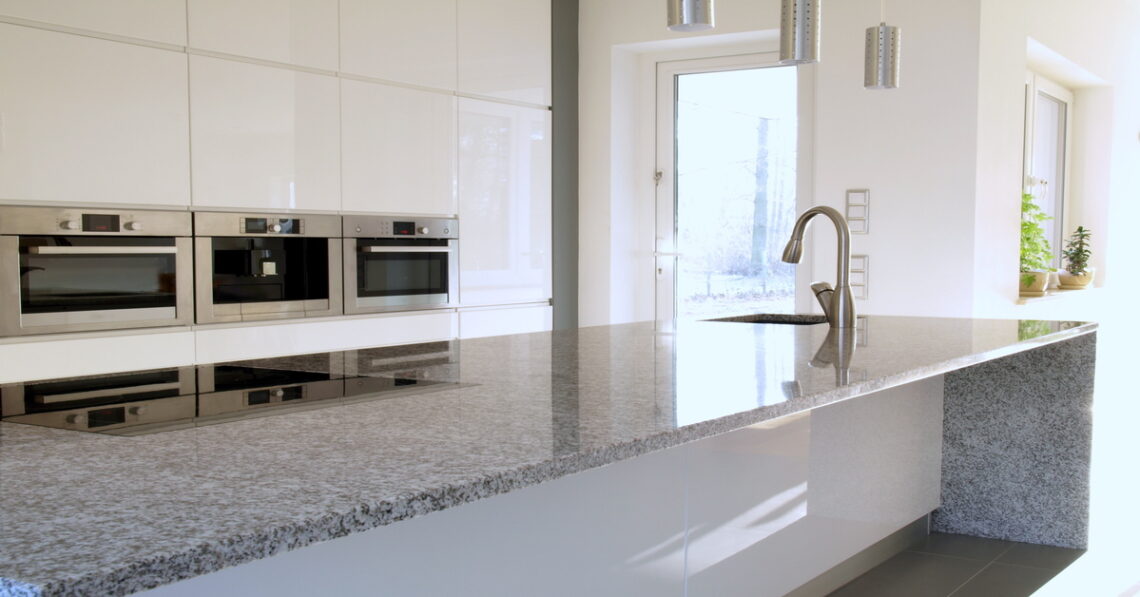 A close-up of a granite kitchen countertop island with a larger kitchen space in the background, and a door.