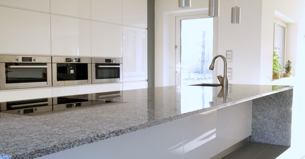 A close-up of a granite kitchen countertop island with a larger kitchen space in the background, and a door.