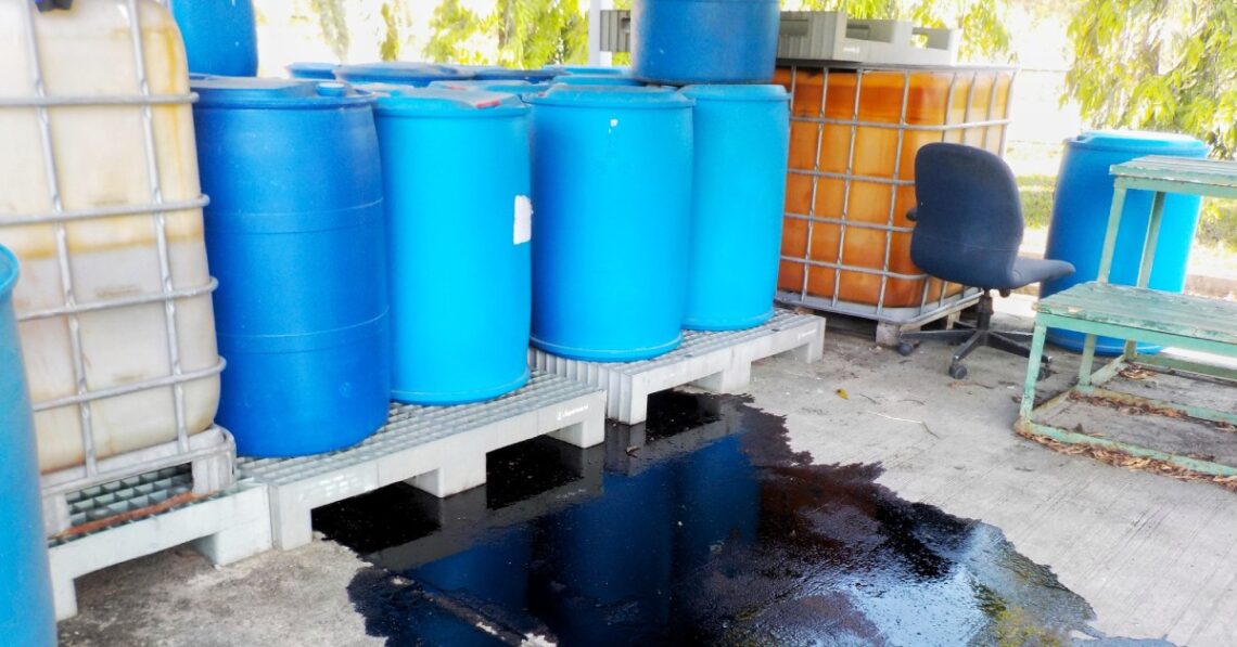 A warehouse with blue drums sitting against the wall and a black spill on the ground surrounding the blue drums.