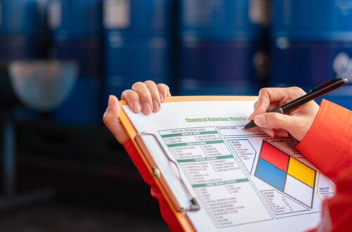 A person holds a clipboard with a hazardous material checklist for chemical handling. Barrels are blurred in the background.