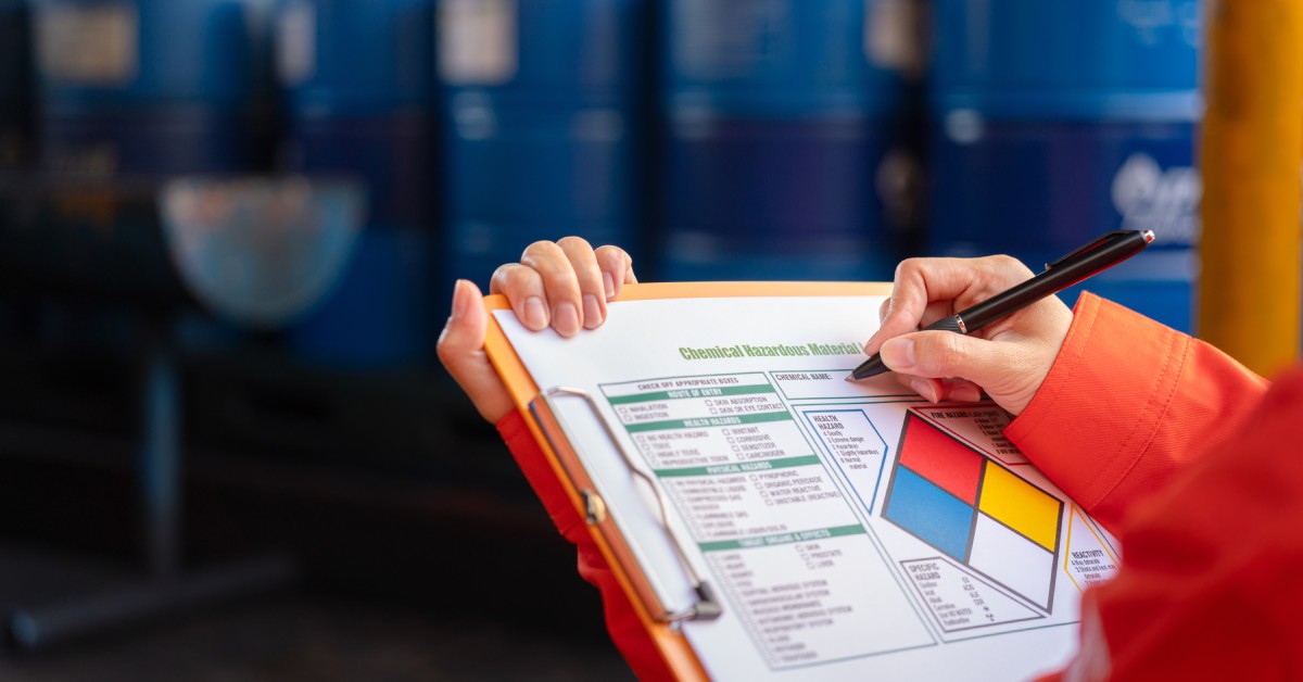A person holds a clipboard with a hazardous material checklist for chemical handling. Barrels are blurred in the background.
