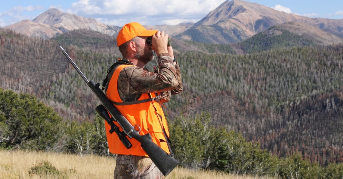 A hunter wearing an orange hat and safety vest looks through some binoculars with his gun strapped to his back.