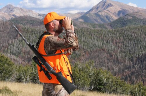 A hunter wearing an orange hat and safety vest looks through some binoculars with his gun strapped to his back.