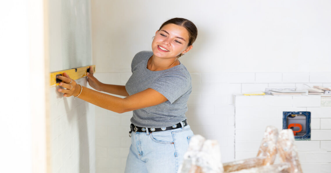 A woman holding a level up against an unfinished wall. There is a ladder covered in white paint in the foreground.