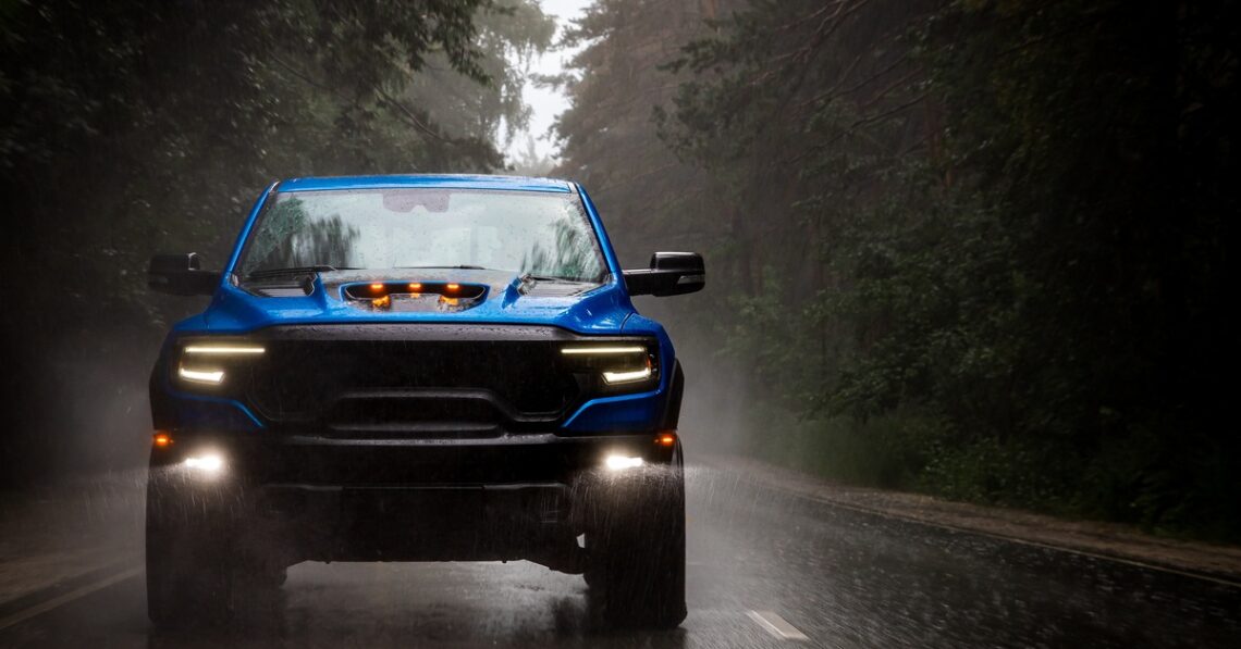 A blue pickup truck drives down a dark, gloomy, foggy road in the rain. The headlights and fog lights are on.