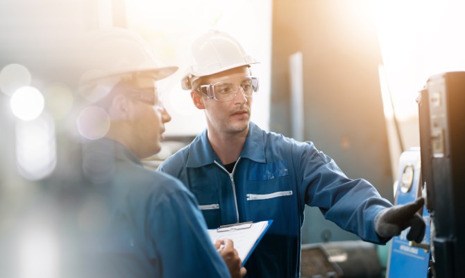 Two engineers standing in front of an industrial machine in the workplace. One employee taps the machine.
